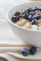 Oatmeal in bowl in close up. Healthy breakfast: Porridge (oatmeal) with banana, blueberry and walnuts in white bowl on linen napkin. Blurred background. Natural food.