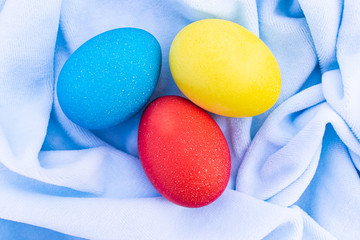 Three multicilired eggs over blue cloth, overhead view. Three bright Easter eggs lie on a crumpled blue cloth. Creative decoration on holiday