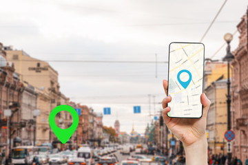 A woman's hand holds a smartphone with an online map app. In the background is a city street with traffic, with a green location icon.Concept of online navigation and GPS