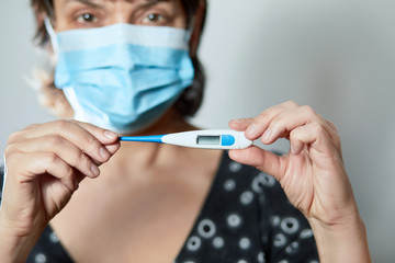 Woman in a mask with Coronavirus reads his temperature on a thermometer. Female measures the temperature. Check body temperature for virus symptoms. Copy space. Coronavirus. 2019 Novel Coronavirus
