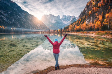 Happy young woman with raised up arms is standing near lake with clear water and mountains at sunset in autumn. Landscape with slim girl, reflection in water, rocks, cloudy sky, orange trees in fall