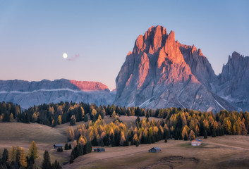 Beautiful mountains with lighted peaks at sunset. Autumn landscape with small wooden houses, mountain valley, meadows with green grass, fall trees, high rocks, sky with moon. Alpe di Siusi in Italy