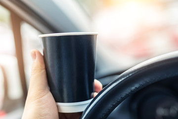 Close-up person hand holding black blank mockup paper cup woth hot coffee or tea takeaway drink driving in urban traffic jam city. Car dashboard and sunrise sunny window background early good morning