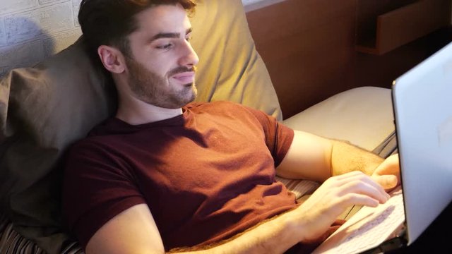 Attractive Young Man with Serious Expression then Smiling, with Laptop on Bed Working on his Start-up Business - Young Male College or University Student Doing Homework, in Bedroom