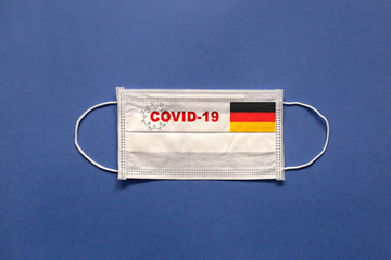 The flag of Germany and inscription COVID-19 on a medical mask on a blue background. Healthcare and medical concept. Pandemic virus COVID-19