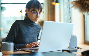 Young business woman in eyeglasses concentrating on screen and typing on laptop while sitting at desk at workplace or cafe. Concept remote work, freelance, using laptop computer.