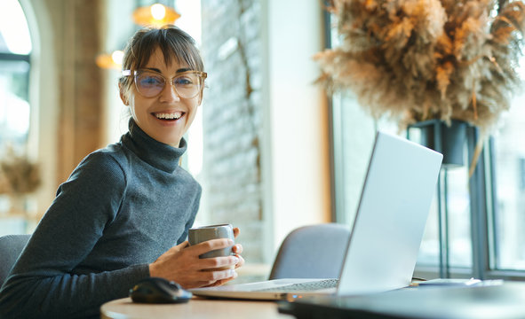 Portrait young woman in glasses drinking coffee, smiling to camera, enjoying studying in coworking space. female sitting in front open laptop computer. Study, learning, remote work, freelance.