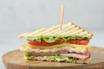 Half a sandwich on a wooden plate. Sandwich filling consists of ham, cheese, lettuce and sliced tomato. Close-up. Light background.