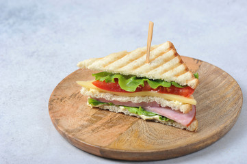 Half a sandwich on a wooden plate. Sandwich filling consists of ham, cheese, lettuce and sliced tomato. Close-up. Light background. Free space for text.