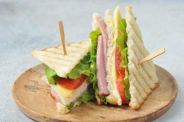 Classic club sandwich on a wooden plate. The sandwich filling includes ham, cheese, lettuce, tomato slices. Light background. Close-up.