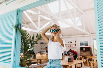 Lovable tanned caucasian woman posing with pleasure near cafeteria. Fashionable fair-haired girl in sunglasses enjoying photoshoot in summer weekend.