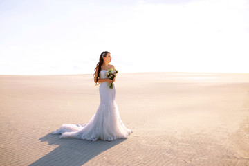 Fototapeta na wymiar Bride in long wedding dress with bouquet stands on sand in desert. Backlight and pastel colors.