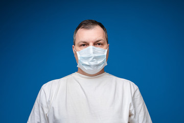 Stock photo portrait of adult man in white t-shirt wearing facial aseptic mask. Isolate on blue background. Studio shot.