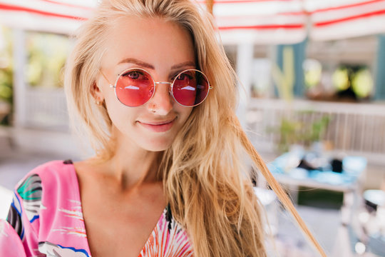 Close-up photo of joyful tanned female model with long blonde hair. Portrait of enthusiastic fair-haired lady in pink sunglasses posing on blur background.