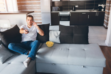 Young man watch tv in his own apartment. Busy guy sitting on couch and talking on phone. Switching...