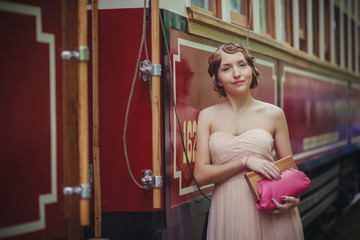 a girl in a light pink retro dress with a vintage hairstyle stands at the rail of the tram