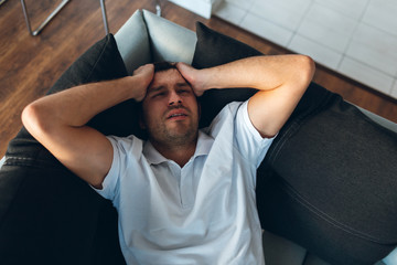 Young man watch tv in his own apartment. Headache, pain or hangover. Hold hands on head and suffer....