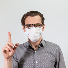 Calm Caucasian man in medical mask and glasses gesturing with forefinger, grey background