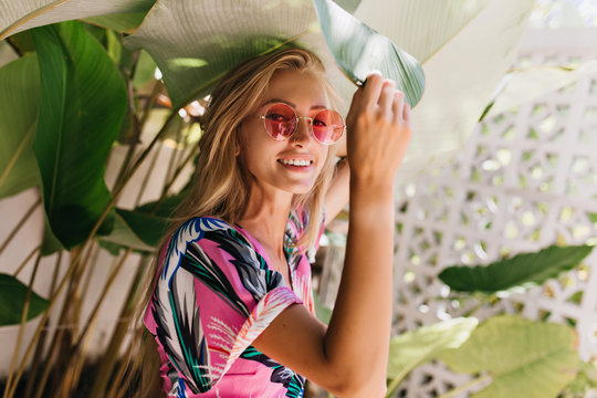 Winsome long-haired woman in pink sunglasses posing beside plants. Outdoor photo of gorgeous young lady in bright attire touching big green leaf.