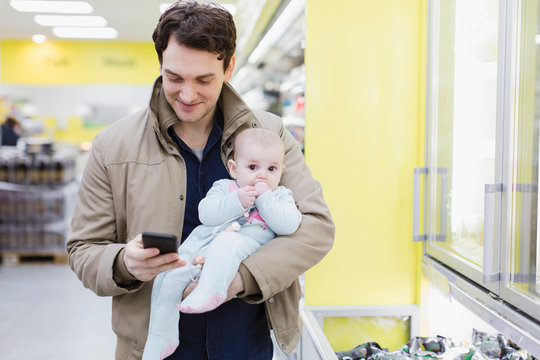 Father With Baby Daughter Checking Smart Phone In Supermarket