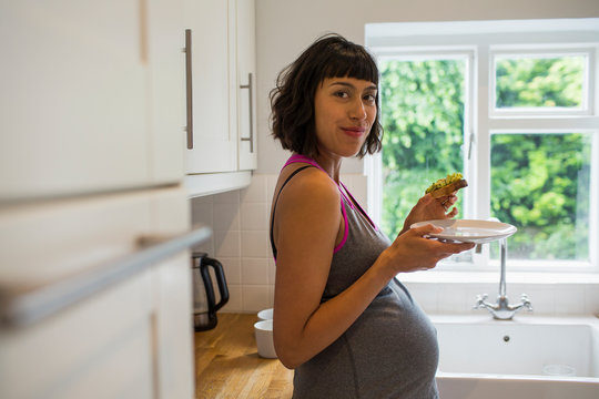 Portrait Confident Pregnant Woman Eating Avocado Toast In Kitchen