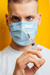 A doctor holds a syringe with a medicinal viral vaccine. Young guy in a white coat in a medical mask on a yellow background. Epidemic pandemic coronavirus concept covid-19. Vertical photo.