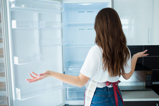 Young Woman On Kitchen During Quarantine. Look At Empty Fridge Shelves With No Food On It. Hungry And Can't Cook. Back View Of Woman Doesn't Know What To Do.