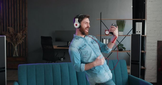 Slow motion of handsome Bearded guy have Fun jumping dancing on bed and listening to Music in Headphones. Man playing imaginary Guitar in Loft Living Room. Playfully Mood on Quarantine.