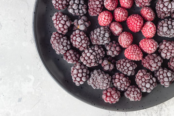 Frozen berries (blackberries and raspberries), covered with hoarfrost on a black ceramic plate. Home harvest. 