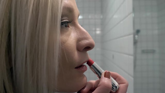 Young blond woman applying red lipstick in hotel bathroom.