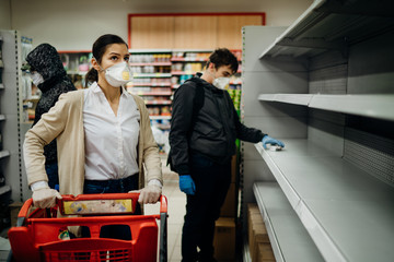 People wearing masks and gloves buying groceries/supplies in supermarket with sold out products.Food supplies shortage.Empty shelves due to coronavirus covid-19 outbreak panic buying and hoarding. - obrazy, fototapety, plakaty