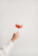 Womans hand in white shirt holding and turning glass of rose wine over white wall background. Wine shop, wine tasting, bar, wine list concept - 334605811