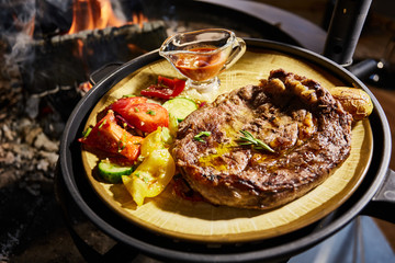 The big piece of the grilled meat and vegetables lies on a plate, Juicy beef steak, near big naked flame, sauce, red coals, a smoke, firewood