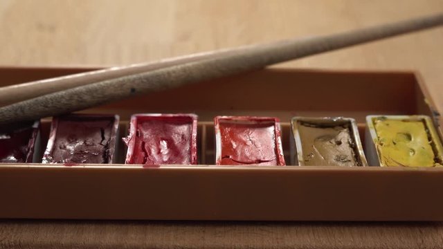 Dolly footage of palette with gouache lies on a wooden table. Different colors of paint on this pallet. The artist paints picture.