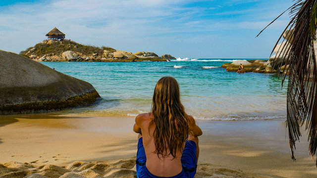 Young man resting on the beach and looking the amazing coast view in Natural National Park Tayrona, Colombia. Back shot of tourist and amazing view of caribbean beach.