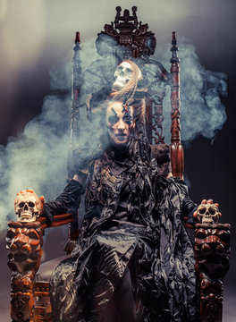 Young beautiful witch sits on a chair. Bright make up, skull, smoke- halloween theme.