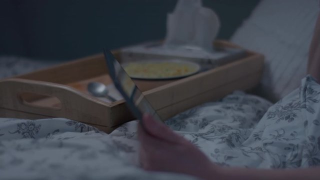 Sick Woman Watches TV On Her Tablet In Bed, Sips Tea From Her Dinner Tray