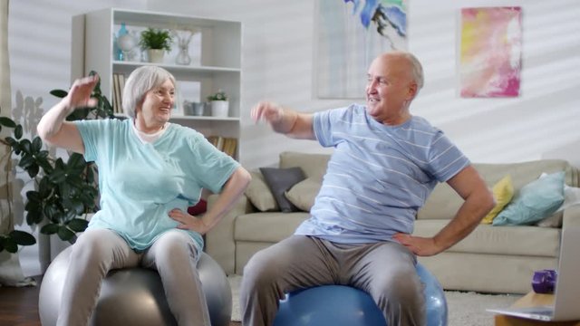 Cheerful senior couple smiling and doing side bend on exercise balls while training together at home