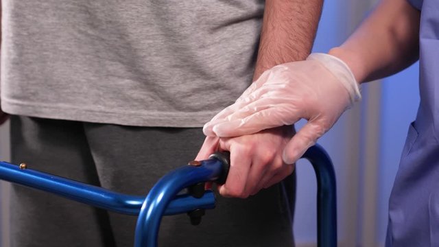 Young man, struggling to use a walking aid, but a gloved hand of a doctor provides reassurance and support to the patient during rehabilitation.