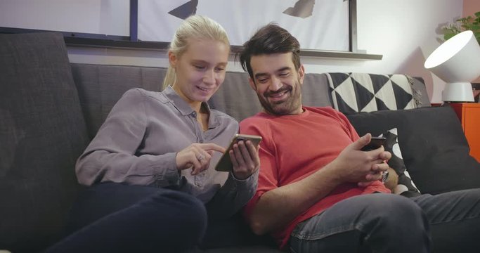 Young positive couple holding smartphones looking at screens watching funny videos, surfing chatting. Social life and networks concept. Caucasian family spend free time at couch at home