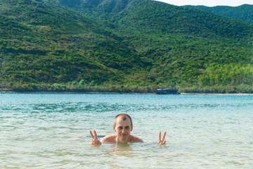 Young man shows peace sign in the water in the sea and green hill
