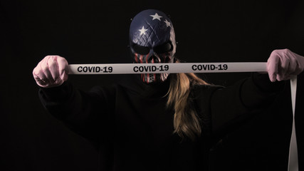 Girl in the scary mask holds a white tape with the inscription covid 19. Isolated on black...