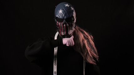Girl in a scary USA color mask and a white ribbon with the inscription covid 19 shows a gesture of death. Isolated on black background. Health care and medical concept. Coronavirus Epidemic