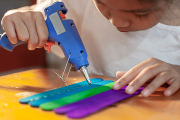 Asian child girl glueing colored ice cream sticks by hot melt electrical glue gun. Children have...