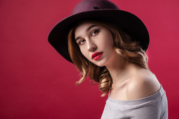 Fashionable attractive Caucasian girl in a large hat to large brim in a silver shiny dress on a red background. Studio retro portrait