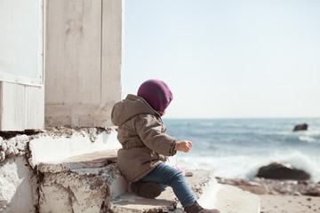 A girl sits on stone steps near a building made of white wooden boards, the baby is dressed in a hat and jacket and looks at the winter sea