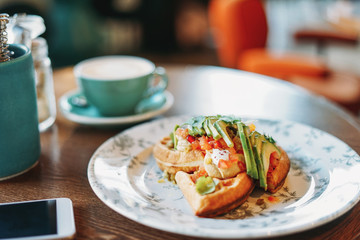 Vegetarian waffles with fresh vegetables and avocado business lunch coffee mobile phone white screen at table in cafe