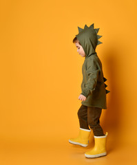 Little brunet kid in khaki dino hoodie with hood and pants, yellow rubber boots. He is walking along orange studio background