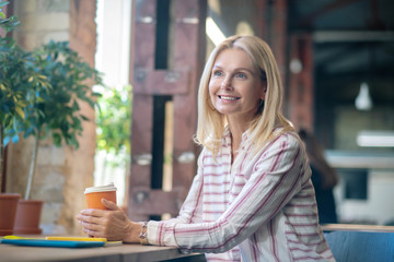 Blonde woman sitting at the window, holding paper cup of coffee