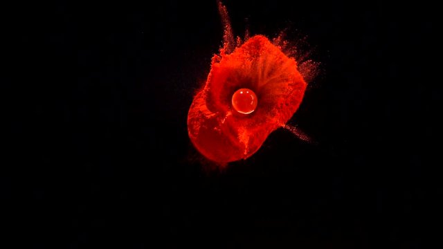 In real slow motion footage against a black background, a clear, water- like ball of gel falls onto the right side of a pile of red powder, bouncing and dispersing a column of red in the air. Top Shot